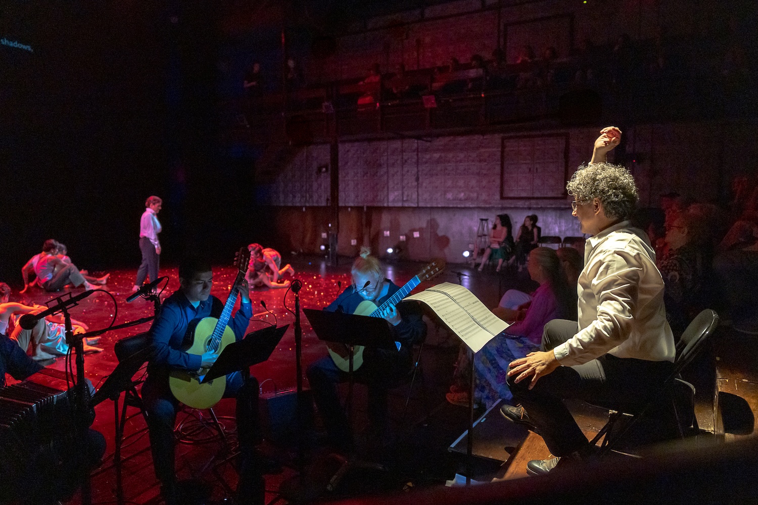 A man with his arm raised directs musicians while dancers perform on stage behind them.