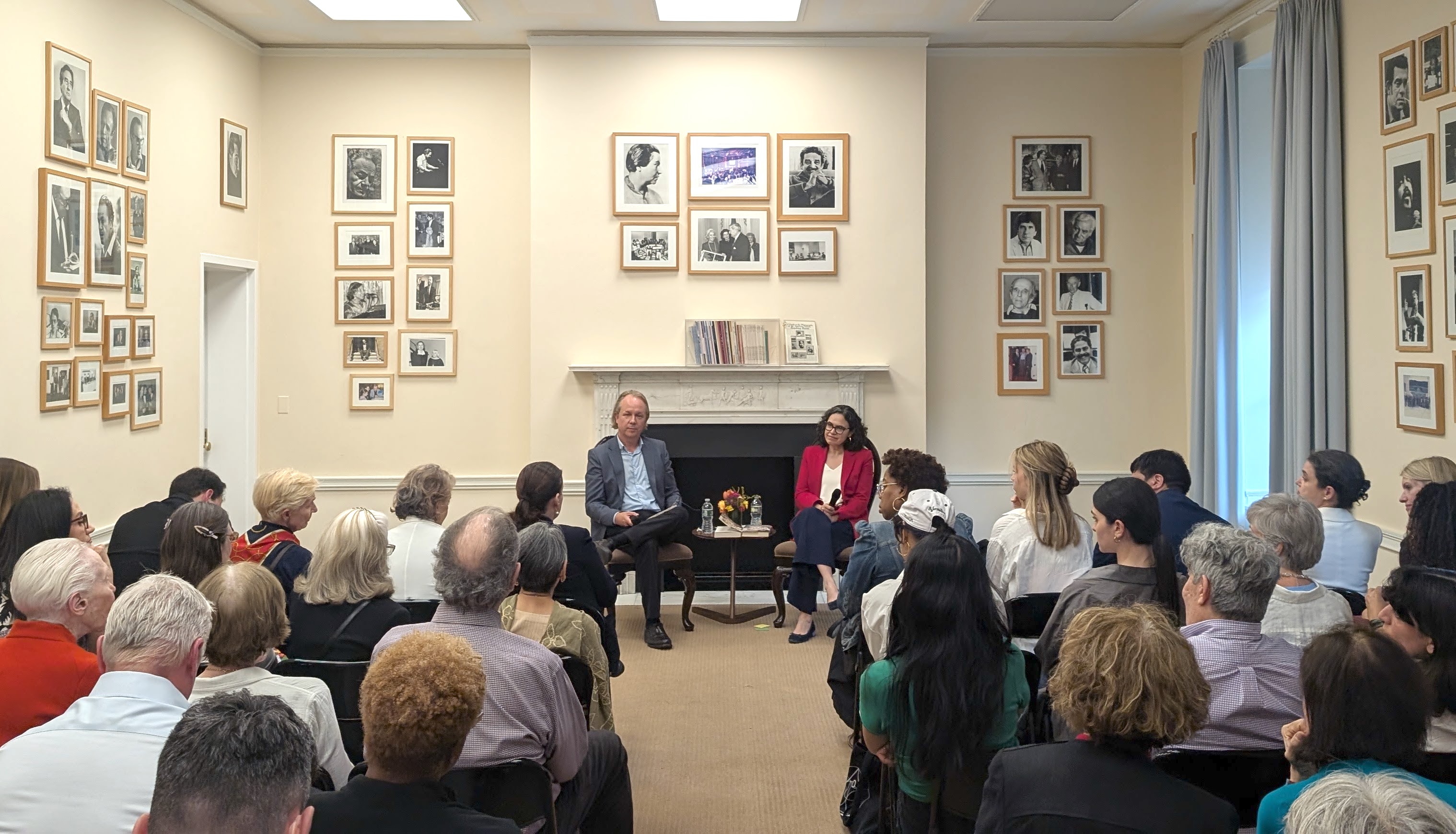 A man and a woman sit in chairs, holding microphones, in front of a large audience in a room surrounded by framed black and white photos of people.