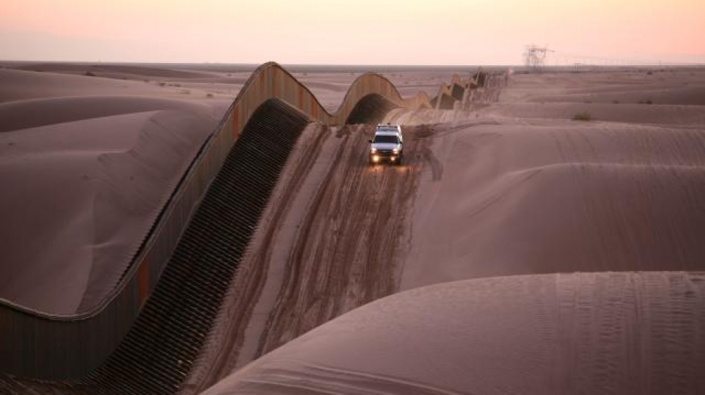 A truck drives over sand dunes alongside a large fence.