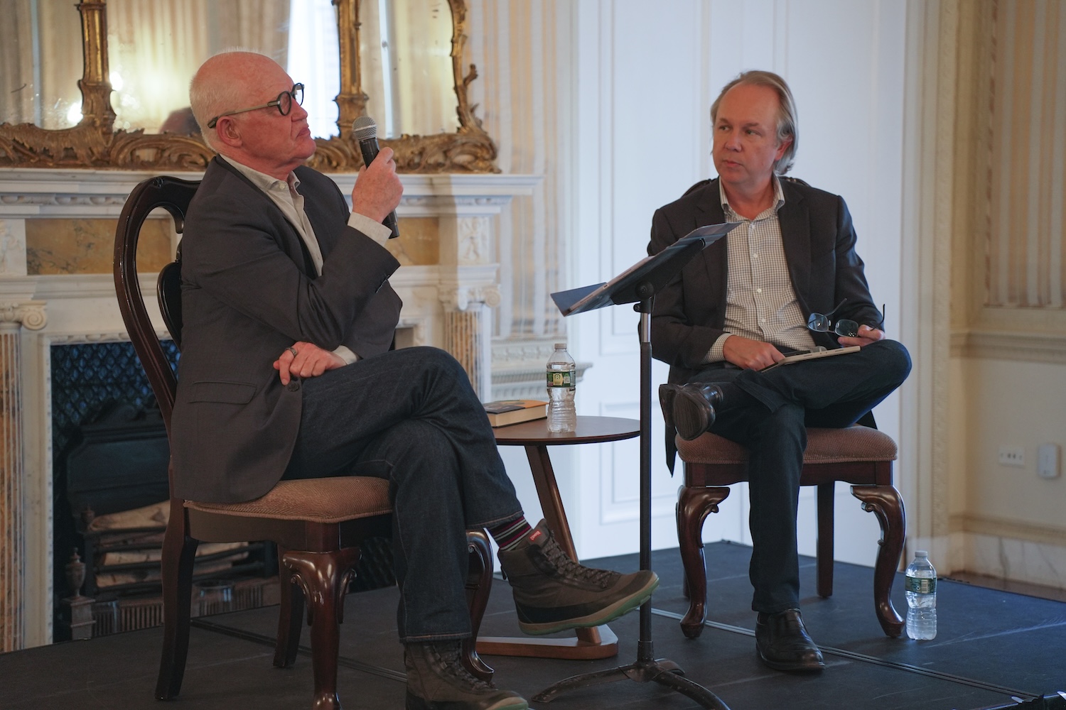 Two men sit in chairs on a stage, one talking into a microphone while the other listens.