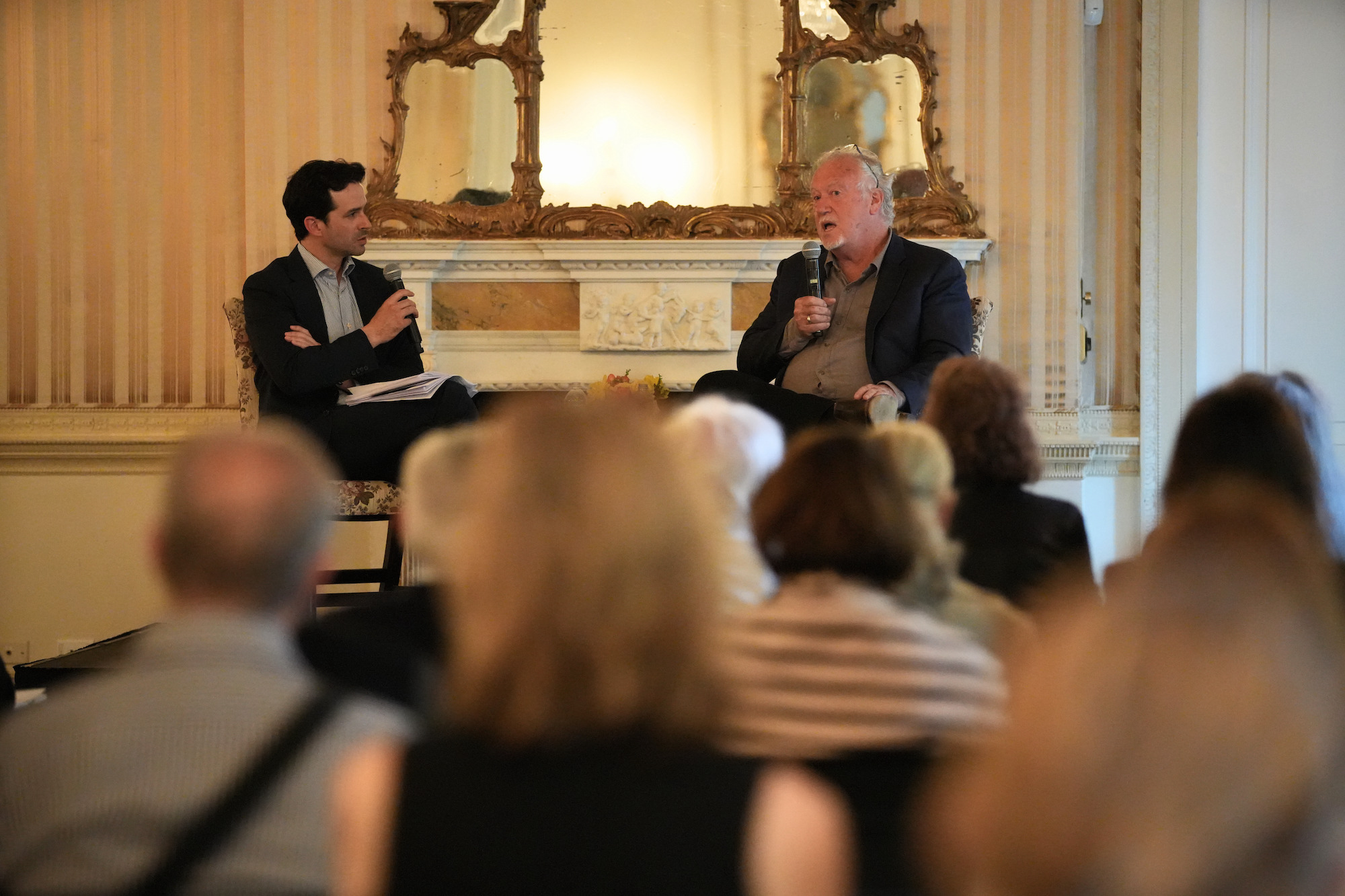 Two men in suit jackets and with microphones sit in front of a seated audience.
