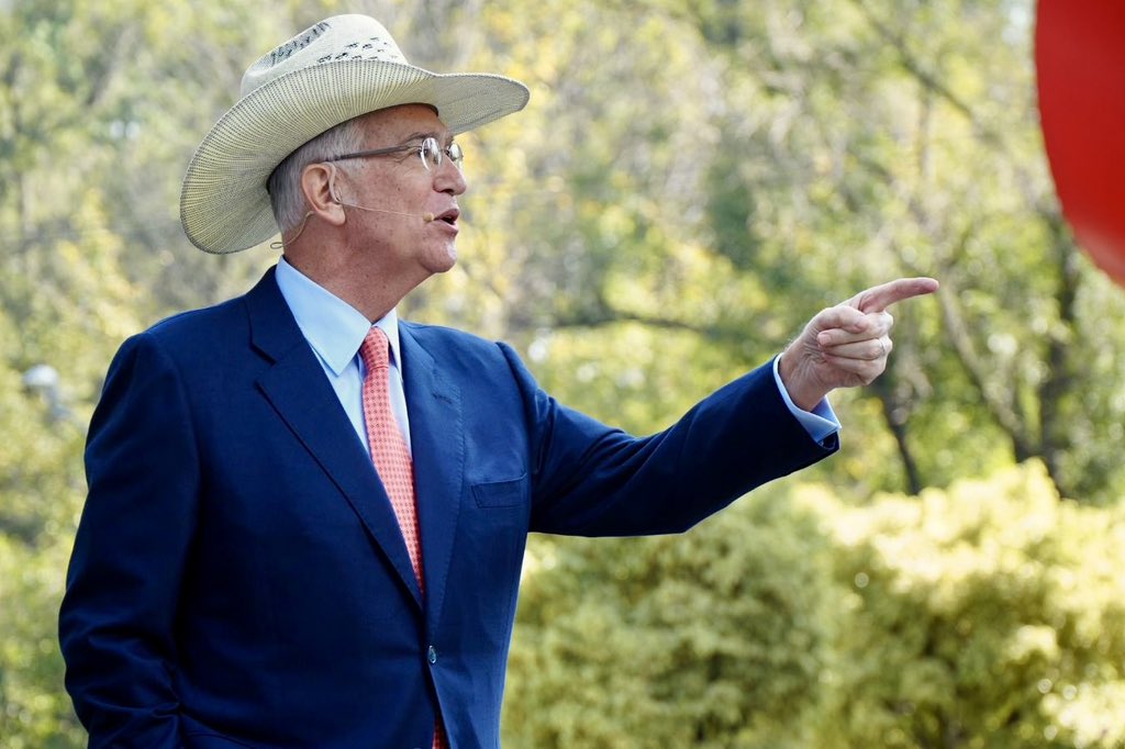 A man in a business suit, glasses, and cowboy hat speaks into a microphone and points against a backdrop of trees.
