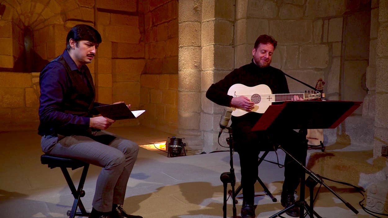 Two men sit on stools in a stone church, one holds a notebook and sings while the other plays a lute.