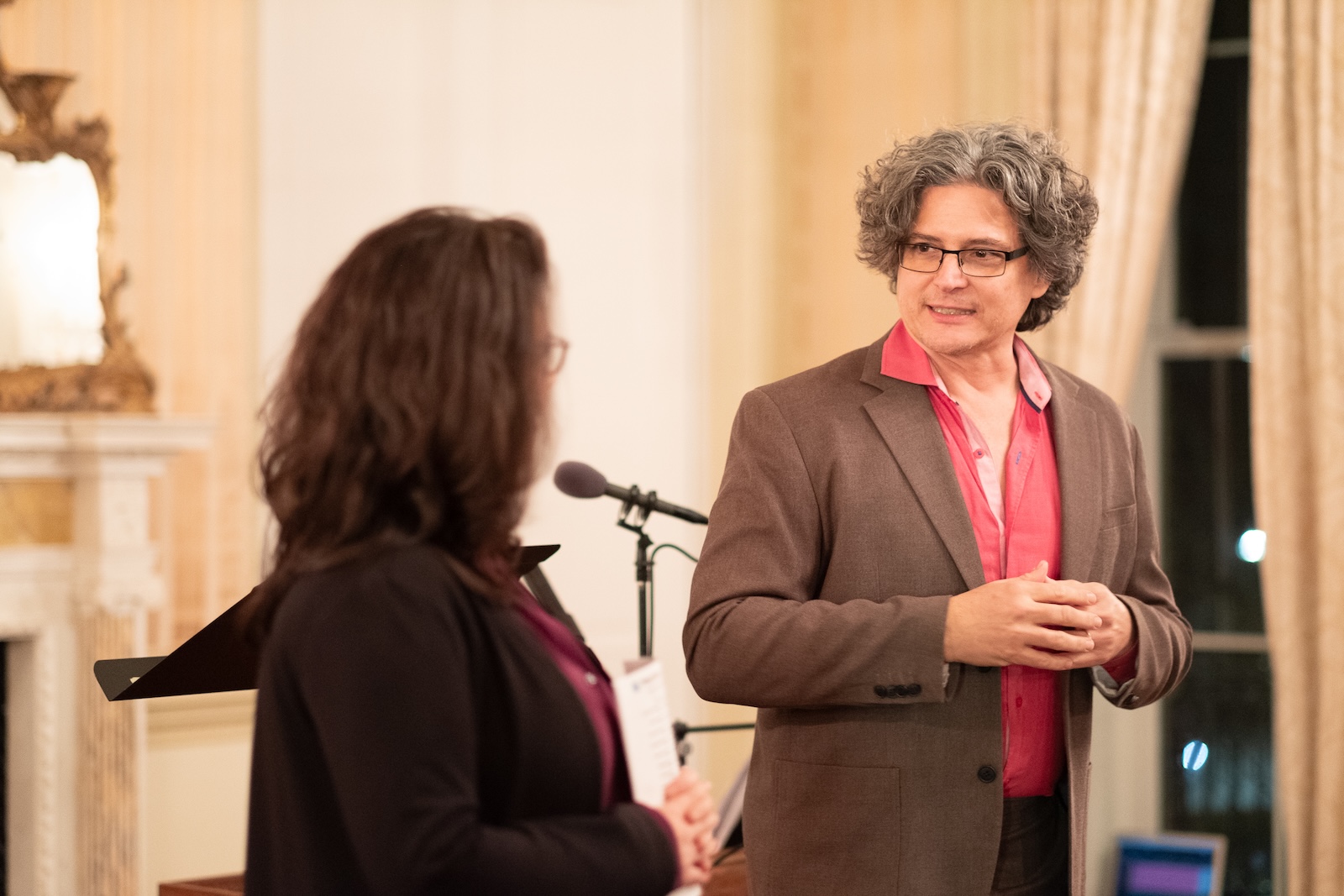 A man with curly hair in a shirt and jacket listens to a woman speaking next to him.