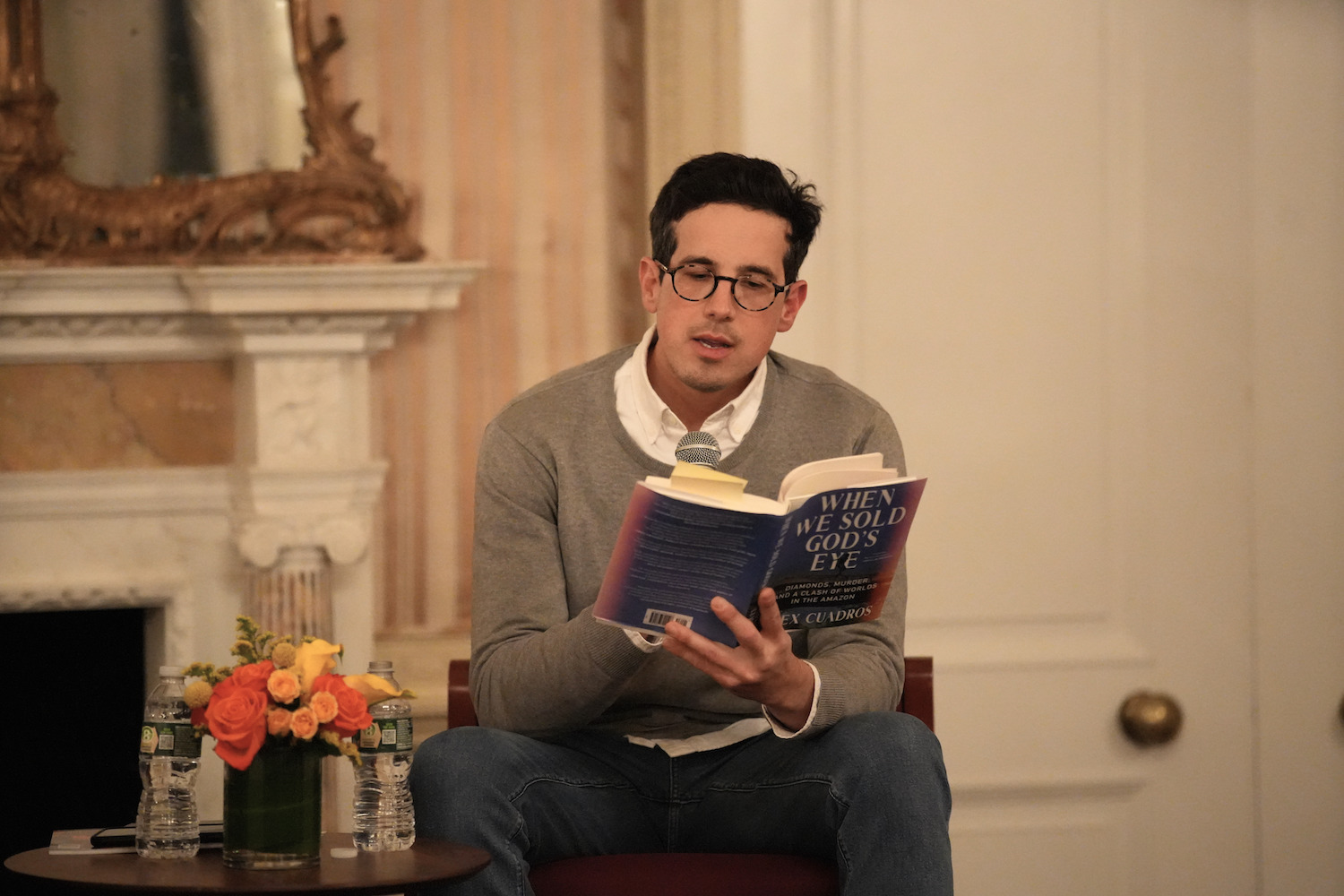 A man with black hair and glasses reads from a book, with flowers beside him, in front of a marble mantlepiece.