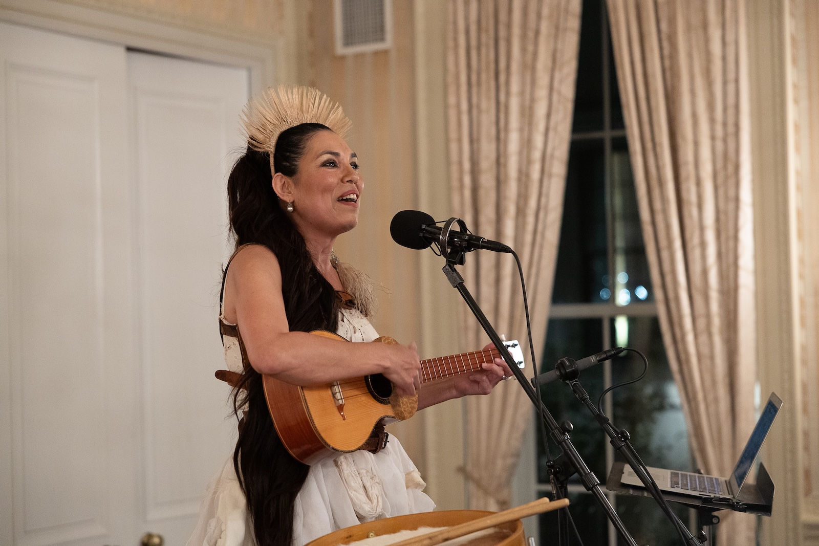 A woman wearing a straw headdress plays a small guitar and sings into a microphone.