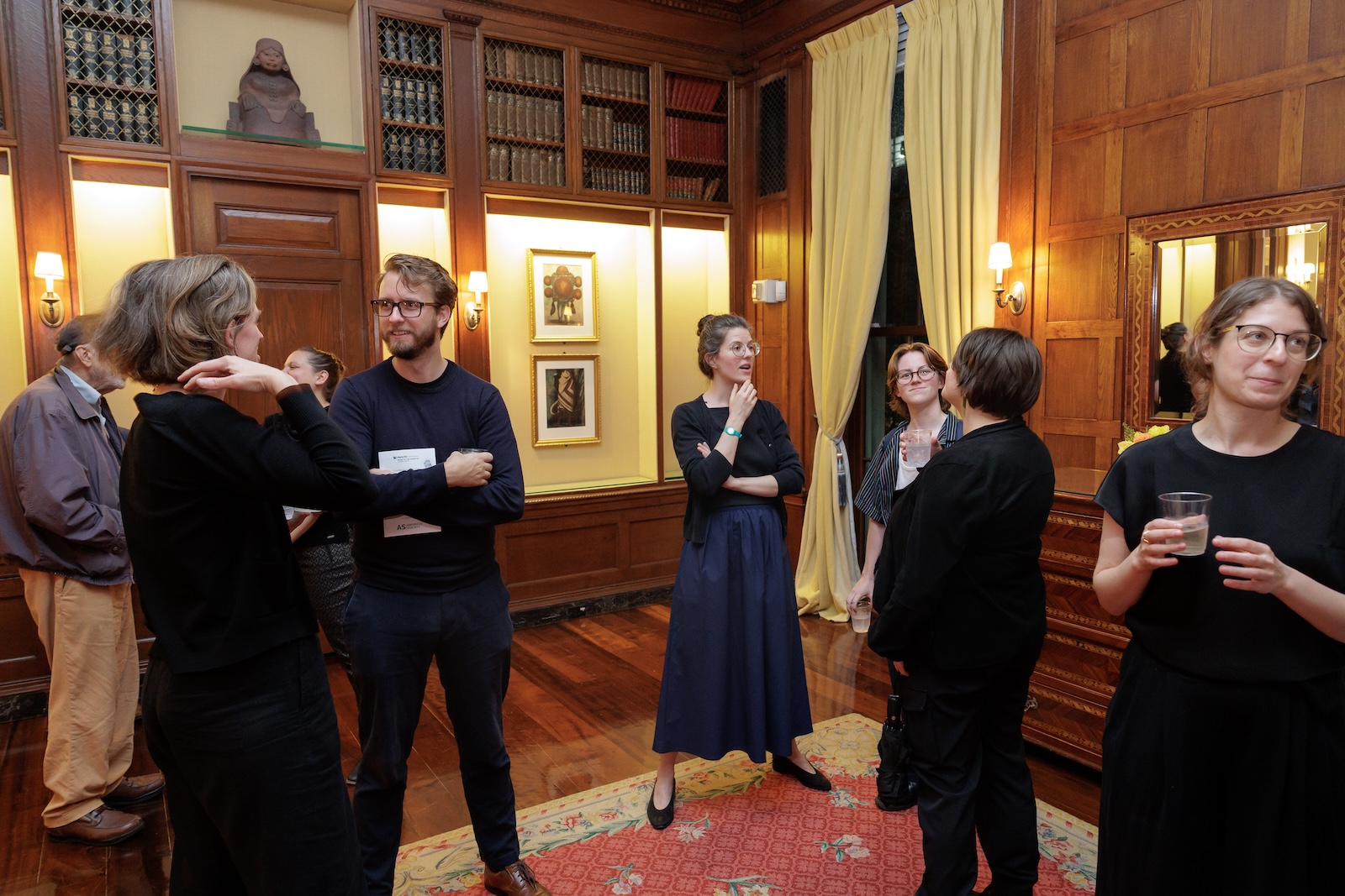 A small crowd mingles in a wood paneled reading room.