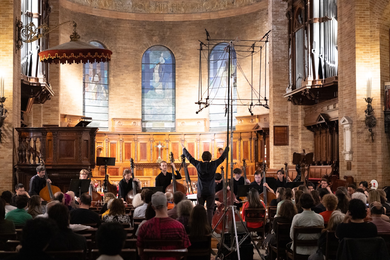 A conductor raises his arms in front of a string orchestra in a chapel with tall stained glass windows in the background.