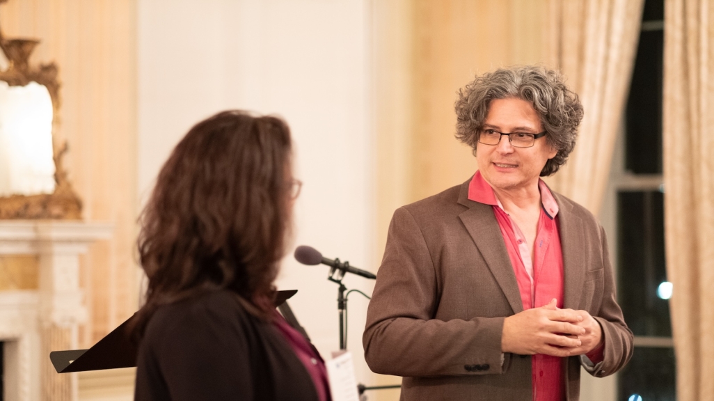 A man with curly hair in a shirt and jacket listens to a woman speaking next to him.