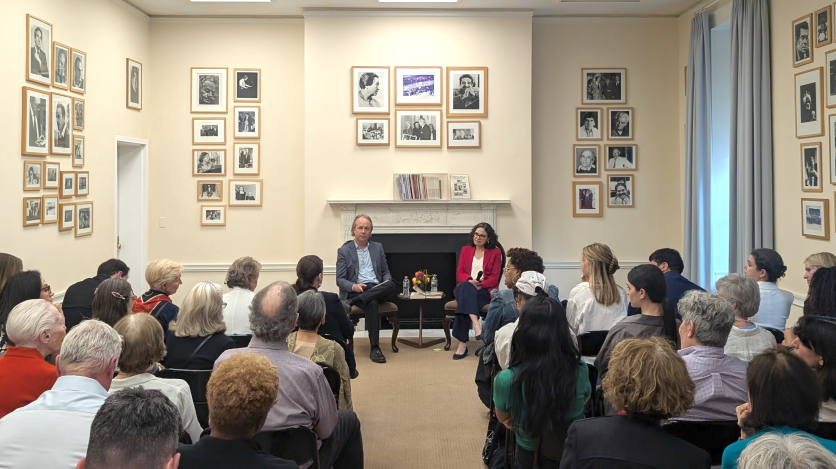 A man and a woman sit in chairs, holding microphones, in front of a large audience in a room surrounded by framed black and white photos of people.