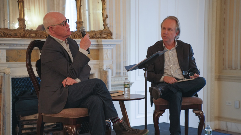 Two men sit in chairs on a stage, one talking into a microphone while the other listens.