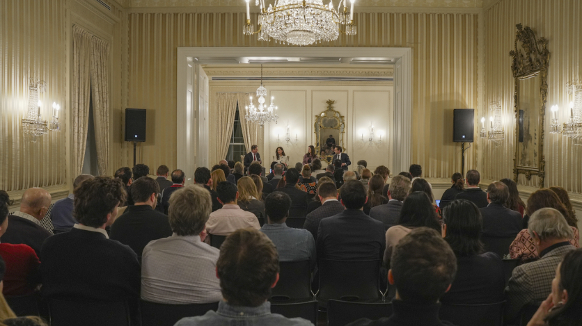 A crowd of 100 or so sits in chairs listening to a panel of two women and two men up on a stage in the front of the room.