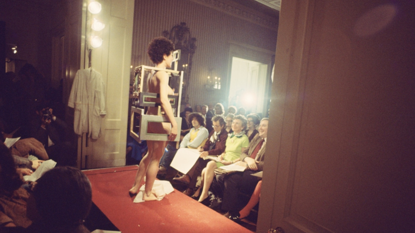 A naked woman with short curly hair stands within what looks like a boxy dress made of matting frames on a runway in front of an audience.