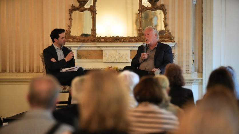 Two men in suit jackets and with microphones sit in front of a seated audience.