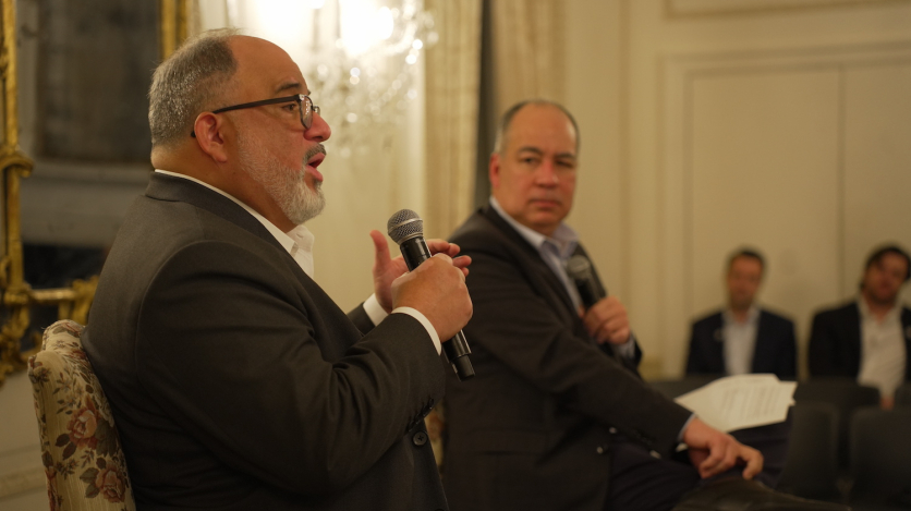 A bearded man seated and in a suit speaks into a microphone while another looks on.