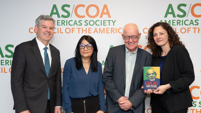 Four people stand, one holding a book, smiling in front of a backdrop.
