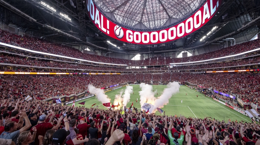 A soccer stadium full of fans cheers with the word "GOOOOOOOOOOAL" on a video screen above the field.