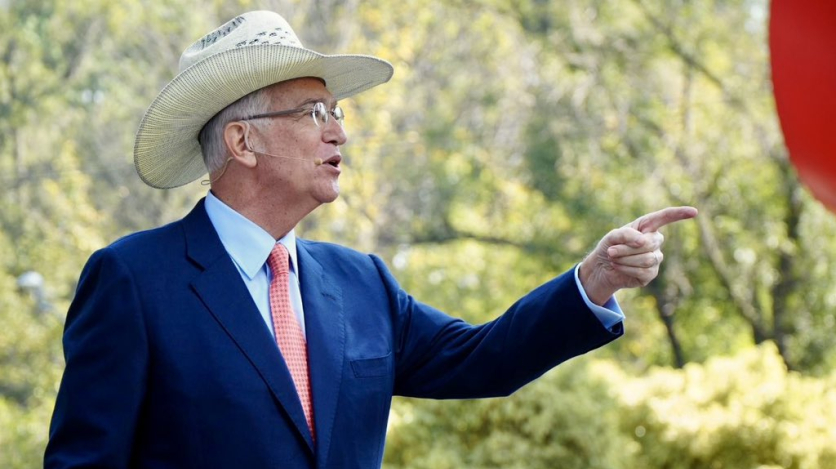 A man in a business suit, glasses, and cowboy hat speaks into a microphone and points against a backdrop of trees.