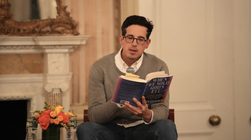 A man with black hair and glasses reads from a book, with flowers beside him, in front of a marble mantlepiece.