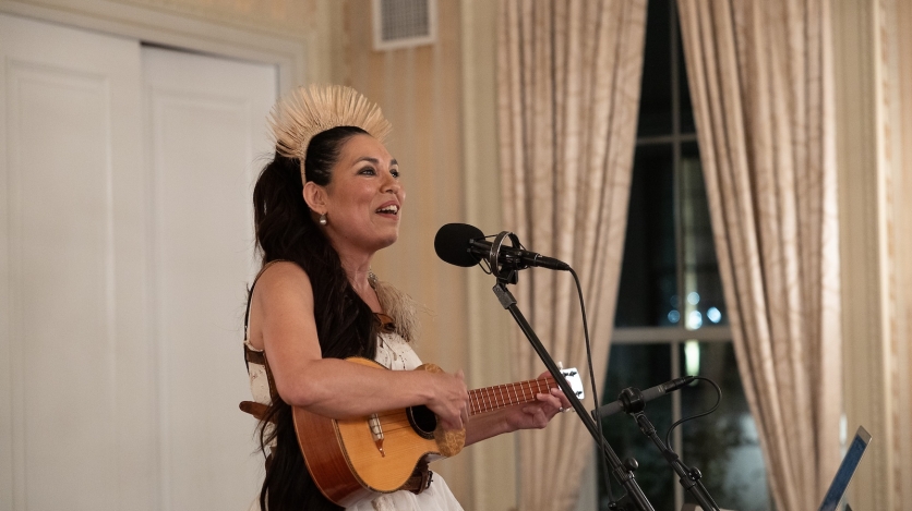 A woman wearing a straw headdress plays a small guitar and sings into a microphone.