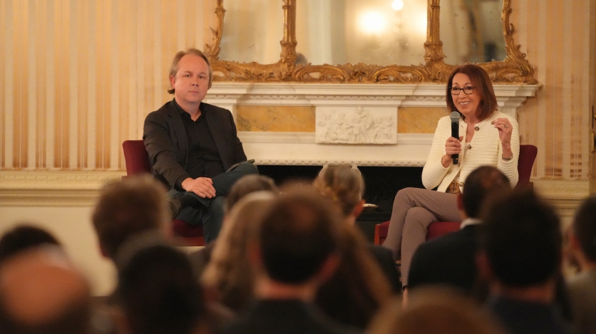 A man and a woman sit in front of an audience with a marble fireplace behind them.