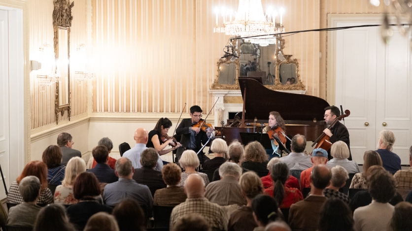 A string quartet plays in front of a small audience in a baroque ballroom.