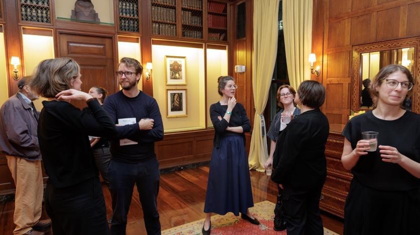 A small crowd mingles in a wood paneled reading room.