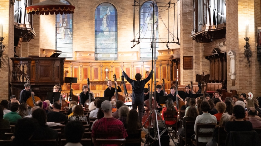 A conductor raises his arms in front of a string orchestra in a chapel with tall stained glass windows in the background.
