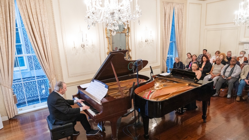 A man and a woman play at two pianos opposite each other in front of an audience in a baroque ballroom.