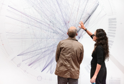 A woman gestures toward a large drawing on a wall while a man looks on.