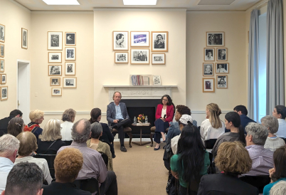A man and a woman sit in chairs, holding microphones, in front of a large audience in a room surrounded by framed black and white photos of people.