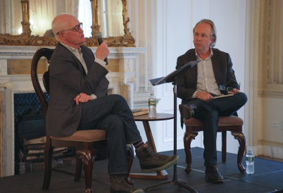 Two men sit in chairs on a stage, one talking into a microphone while the other listens.