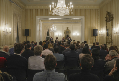 A crowd of 100 or so sits in chairs listening to a panel of two women and two men up on a stage in the front of the room.