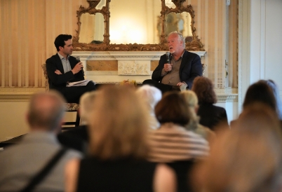 Two men in suit jackets and with microphones sit in front of a seated audience.