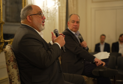 A bearded man seated and in a suit speaks into a microphone while another looks on.