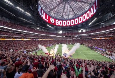 A soccer stadium full of fans cheers with the word "GOOOOOOOOOOAL" on a video screen above the field.