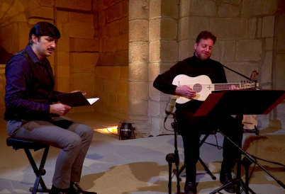 Two men sit on stools in a stone church, one holds a notebook and sings while the other plays a lute.