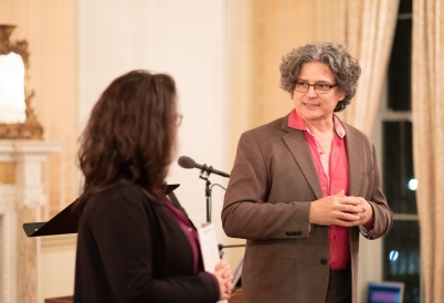 A man with curly hair in a shirt and jacket listens to a woman speaking next to him.