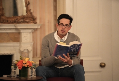 A man with black hair and glasses reads from a book, with flowers beside him, in front of a marble mantlepiece.