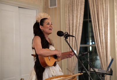 A woman wearing a straw headdress plays a small guitar and sings into a microphone.