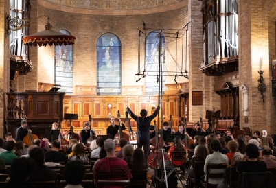 A conductor raises his arms in front of a string orchestra in a chapel with tall stained glass windows in the background.