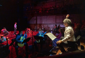 A man with his arm raised directs musicians while dancers perform on stage behind them.