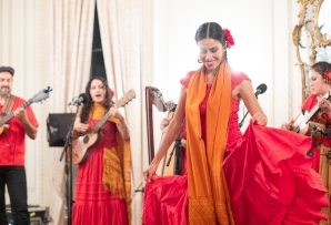 A woman in a red dress and orange shawl dances in front of musicians dressed similarly.