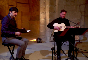 Two men sit on stools in a stone church, one holds a notebook and sings while the other plays a lute.