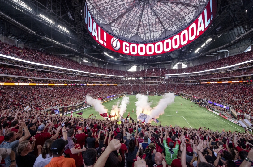 A soccer stadium full of fans cheers with the word "GOOOOOOOOOOAL" on a video screen above the field.