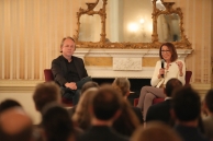 A man and a woman sit in front of an audience with a marble fireplace behind them.
