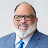 Juan Cruz in a blue pinstripe suit and bright blue tie wears glasses and has a full white beard.