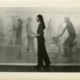 A woman with long dark hair faces left and walks in front of a silhouetted print of people.