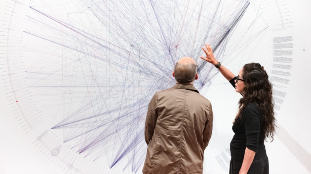 A woman gestures toward a large drawing on a wall while a man looks on.