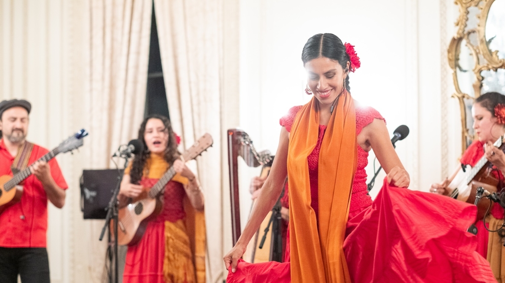 A woman in a red dress and orange shawl dances in front of musicians dressed similarly.